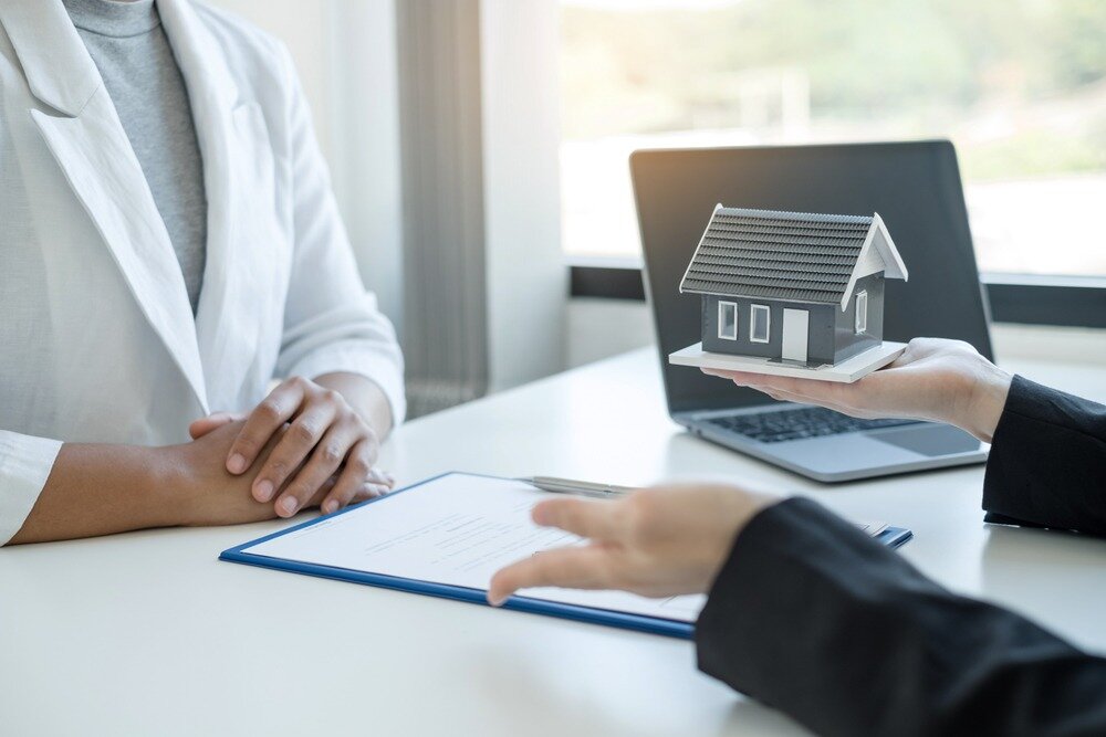 A hand holds a model home up while another person sits on the other side of the desk with folded hands.