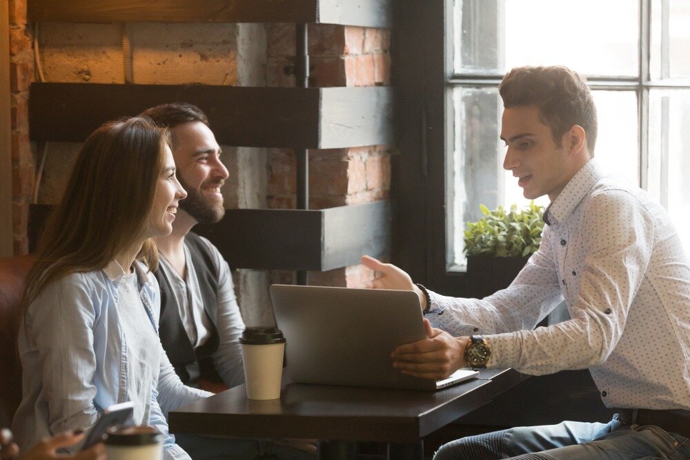 A young man in a button up shirt sits across a desk from a smiling couple and gestures at a laptop that he’s turning their way.