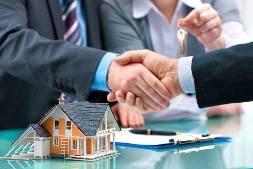 Two people wearing suits shake hands over a table that has a clipboard and an orange model home on it while another hand in the background holds a set of keys.