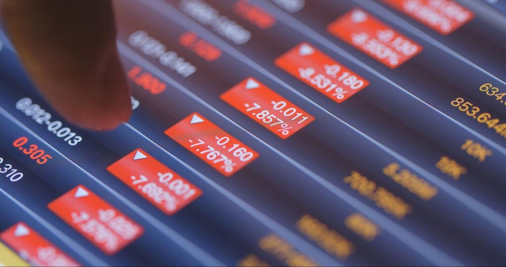 A finger points at red tabs on a tablet that indicates stocks are trending downwards.