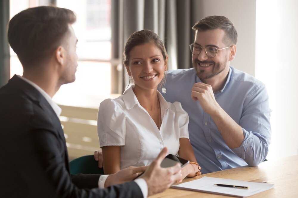 A young man in a black blazer holds an electronic tablet while speaking to a woman and man who are both smiling.