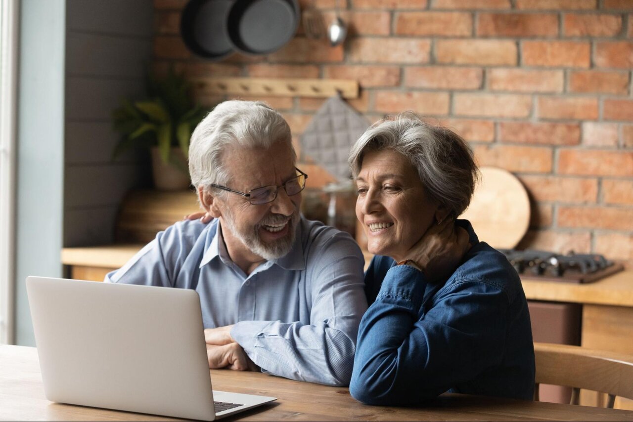 A smiling older couple sitting in their kitchen and looking at their laptop.