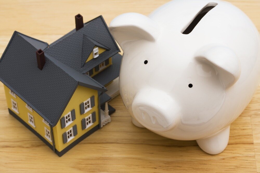 A model of a yellow home with a gray roof and trim is situated next to a white piggy bank.