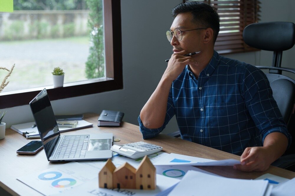 An Asian man in a blue button-up sits at a desk cluttered with documents, a calculator, a model home, and a laptop.