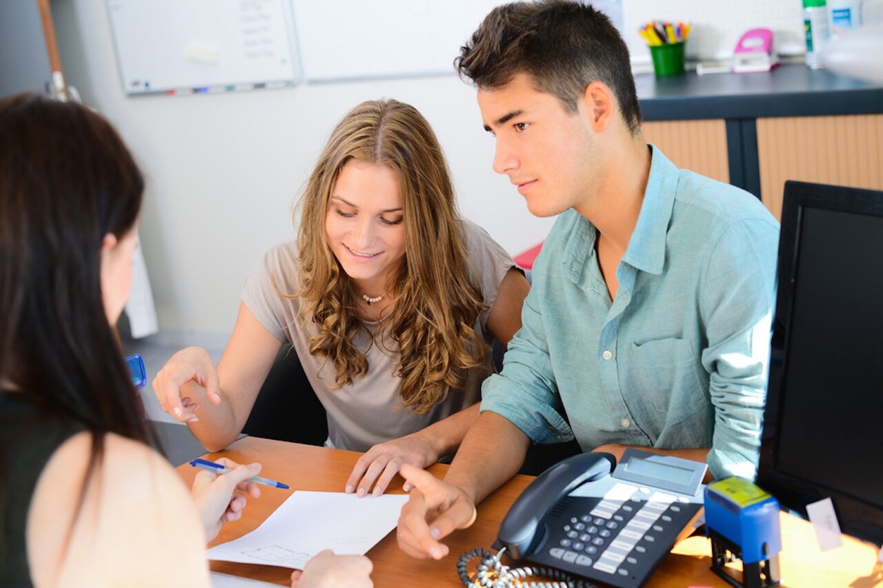 A young white man and woman sitting across the desk from a woman holding out a document and going over it with a pen.