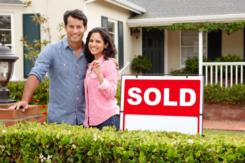 A smiling man and woman holding up a set of keys stand in front of a house next to a red “SOLD” sign.