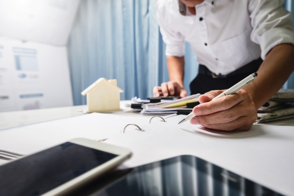 A person in a white button-up uses a pen to write something in a binder that’s on top of a desk cluttered with a phone, a tablet, a model home, and other paperwork.