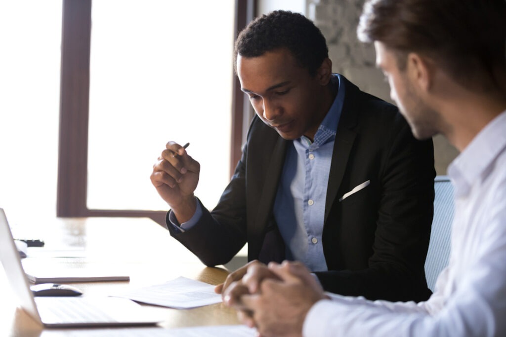 Image of a Black man reviewing documents sitting next to a white man at a table with a laptop on it.