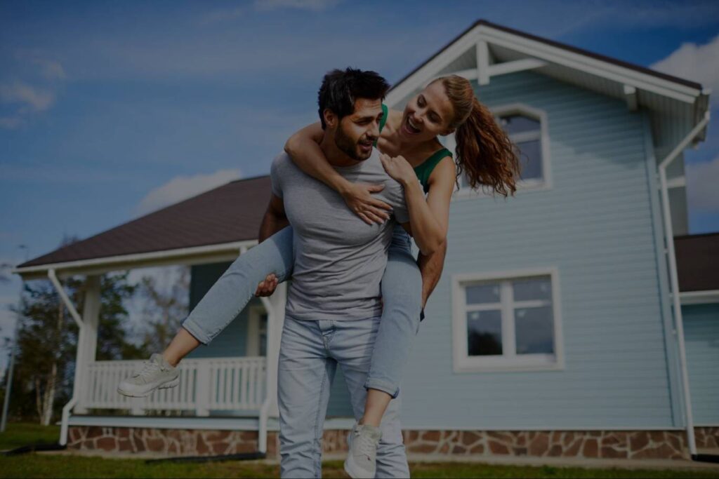 A man giving his wife a piggyback ride in front of the home they just purchased.