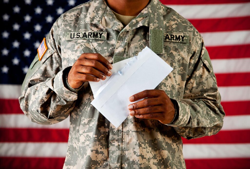 Man in military uniform standing in front of an American flag and opening a letter.