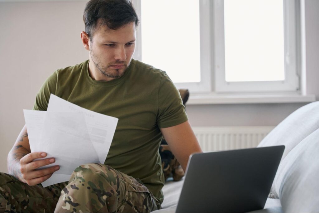 Man in military uniform holding paperwork and typing on a laptop.