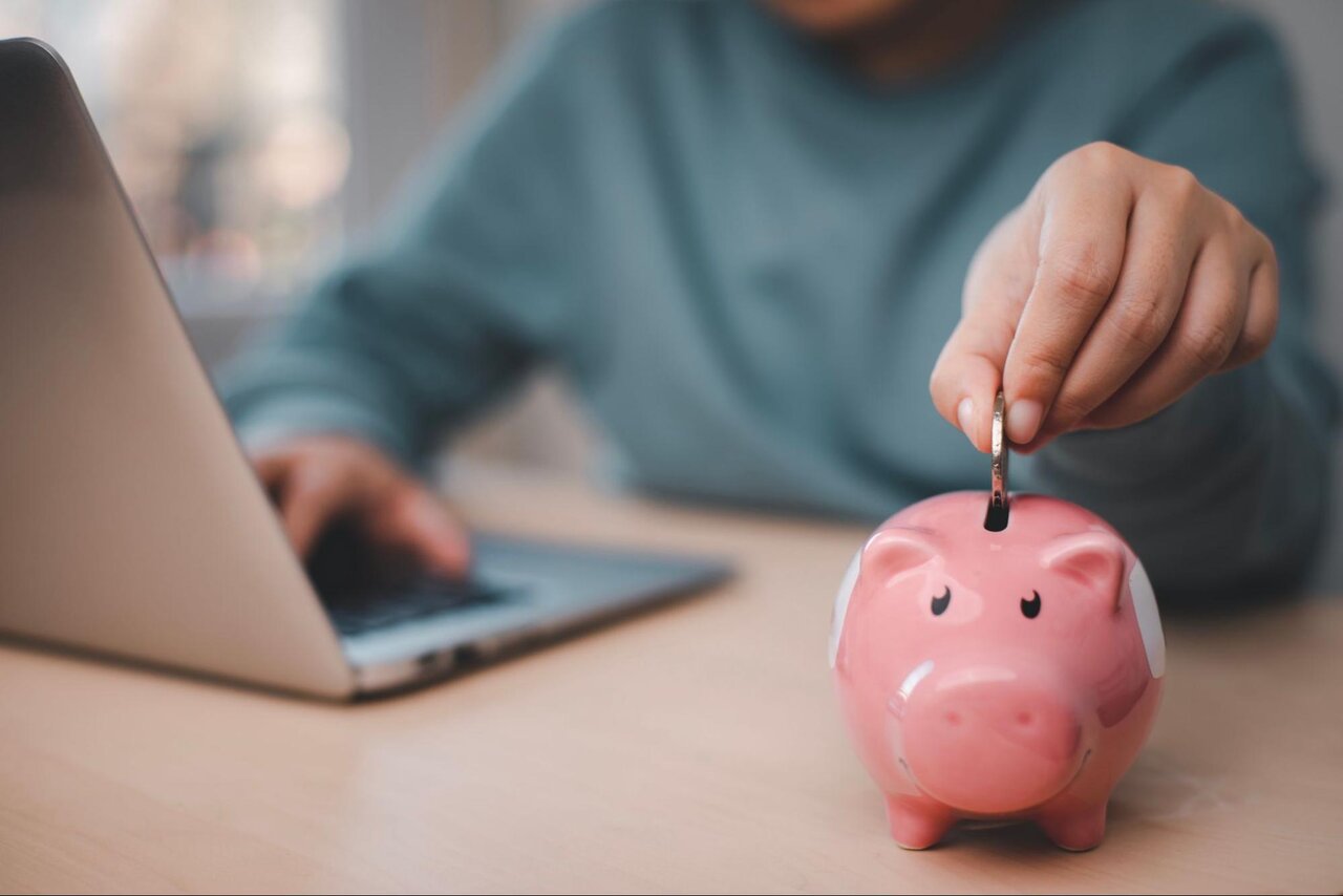 A person working on their laptop places a coin into a pink piggy bank.