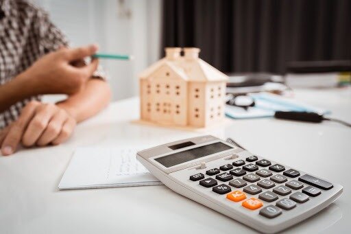 A calculator in the foreground with a house figurine in the background out of focus.