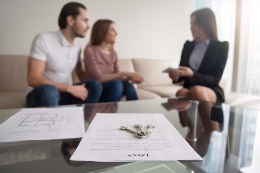 A photo of keys, loan documents and house blueprints in focus in the foreground, with a couple and a lender out of focus in the background.