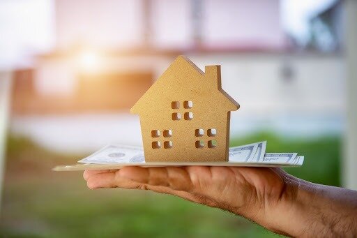 A hand holding a stack of money with a small house figure on top.