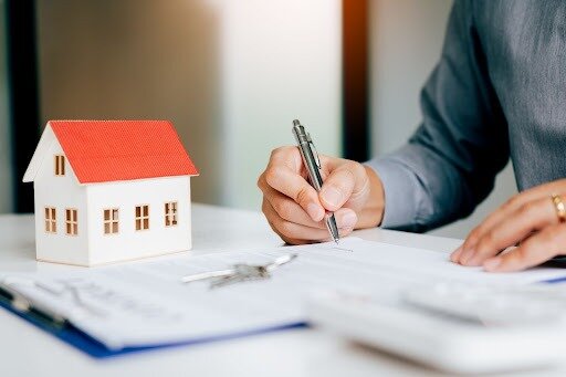 A photo with documents blurred in the foreground on a table with keys and a tabletop house and a person holding a pen to the papers.