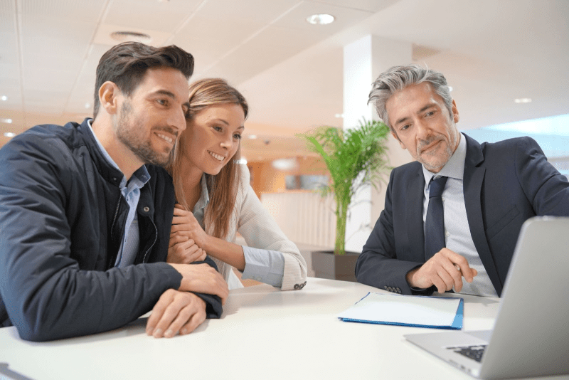 A mortgage lender in a black suit shows a smiling young couple something on his laptop.