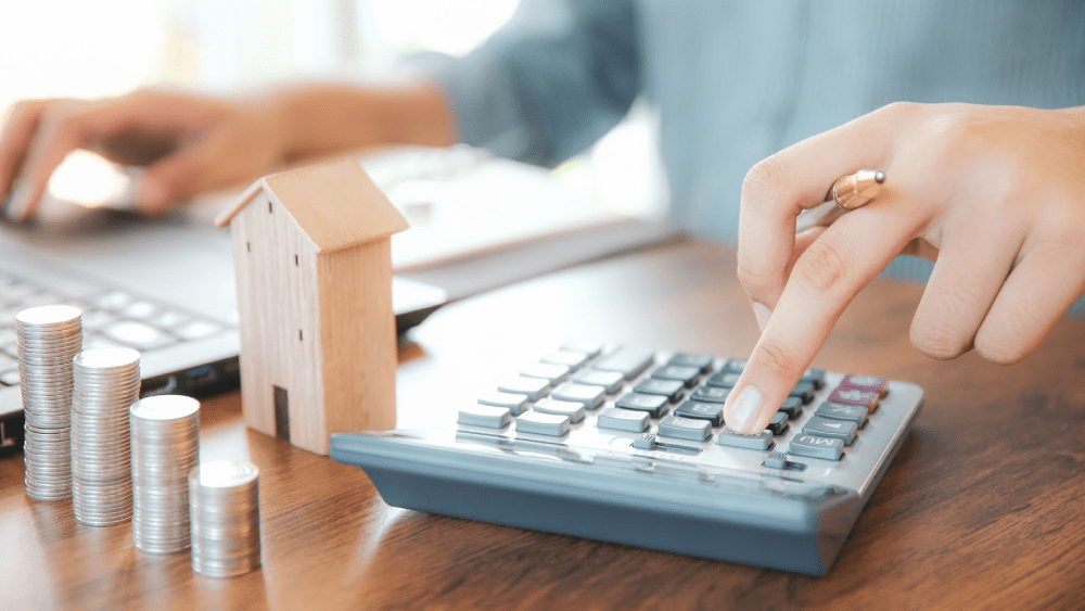 A person types on a calculator near stacks of coins and a model home.