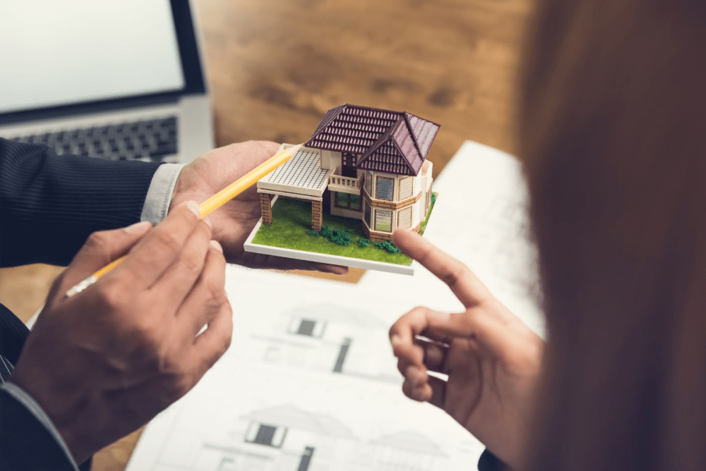 A person gestures towards a small model home using a pencil.