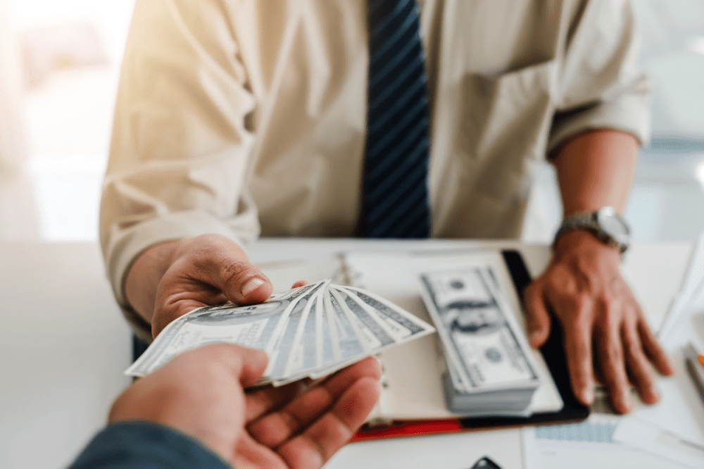 One person hands a stack of cash to a man in a collared shirt and tie.