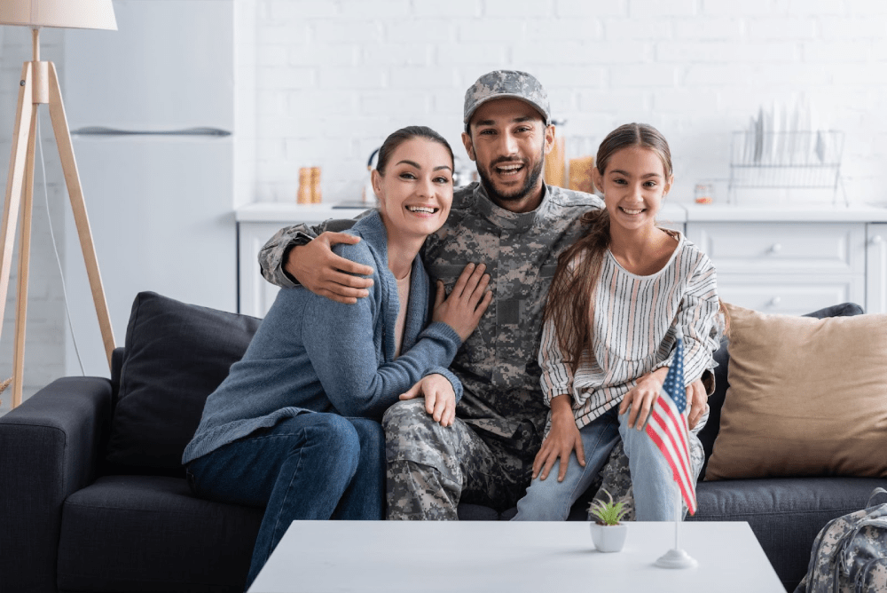 A happy veteran in camo sits with his arms around his wife and daughter.