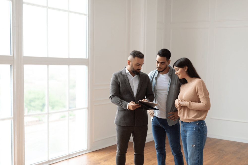 A realtor wearing a dark suit shows a young couple documents in an apartment he’s showing.