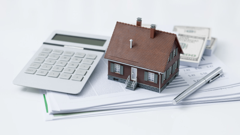 A model home sits on top of a report and next to a calculator and stack of money.