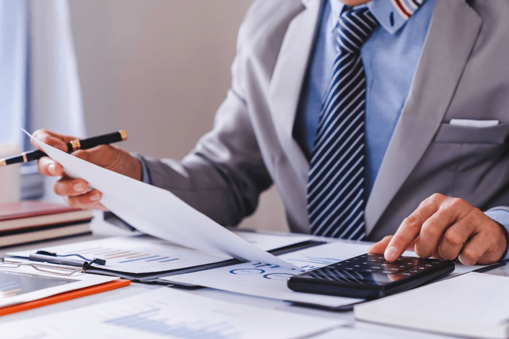 A man in a suit reviews reports and graphs while using a calculator.