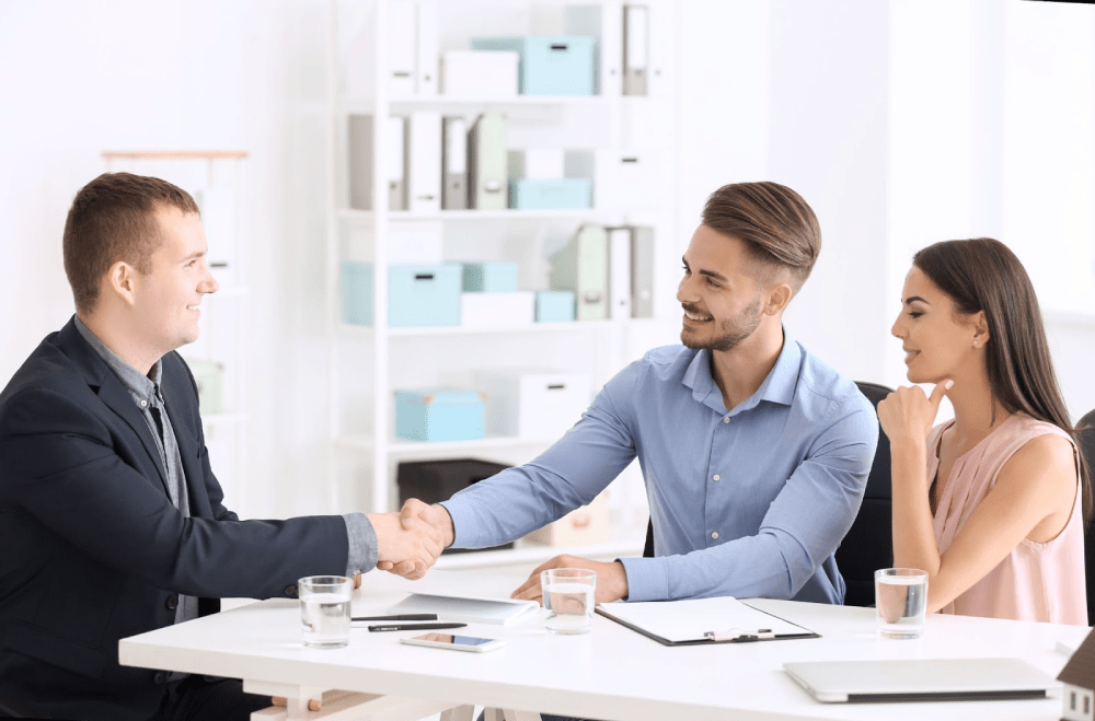 A young couple shakes hands with their lender in his white office.