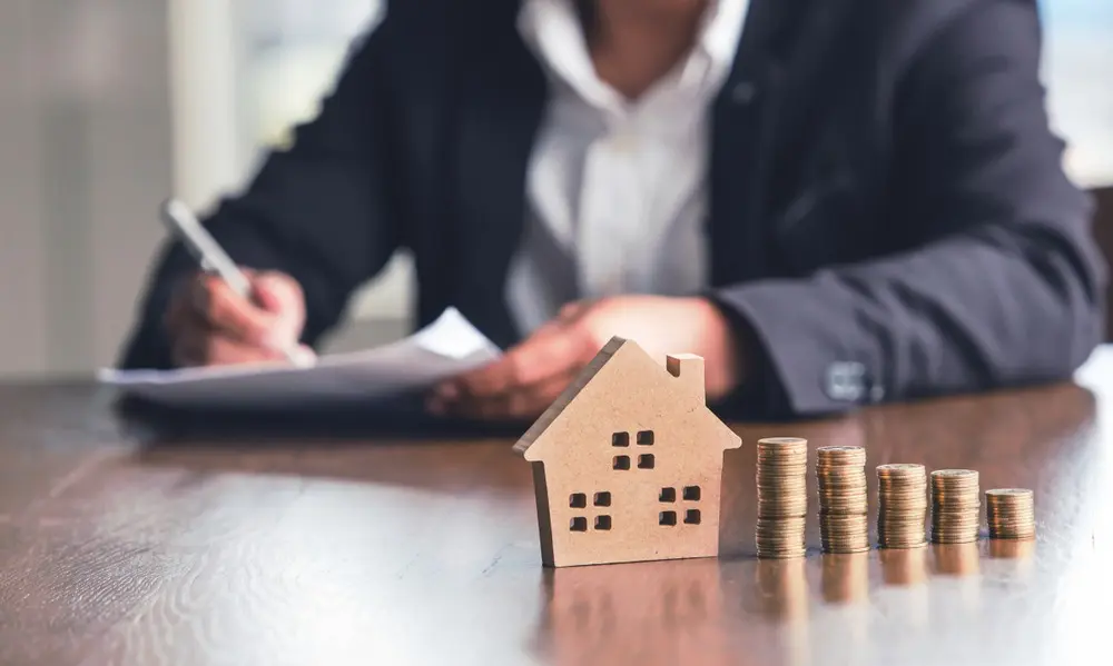 A model home sits next to five stacks of coins while, in the background, a person in a suit writes on a document.