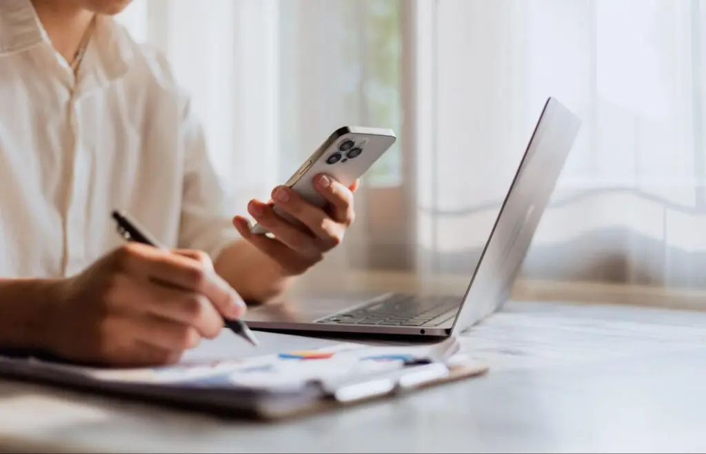 Image of a man using his phone and writing something down with a laptop in front of him.