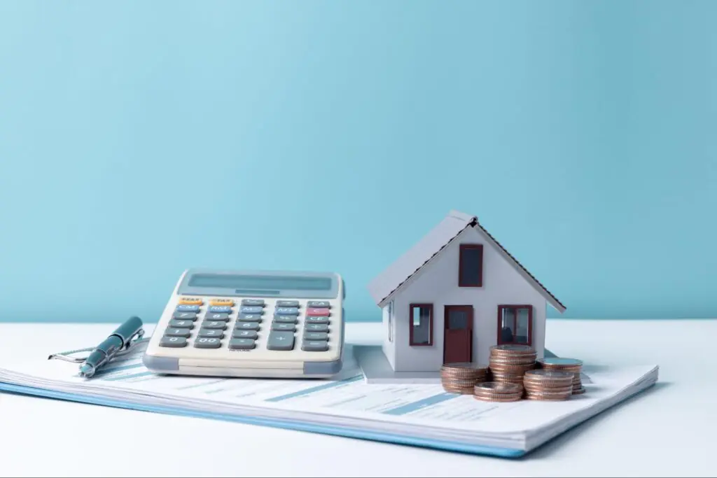 A grey model home positioned by a stack of coins and a calculator.