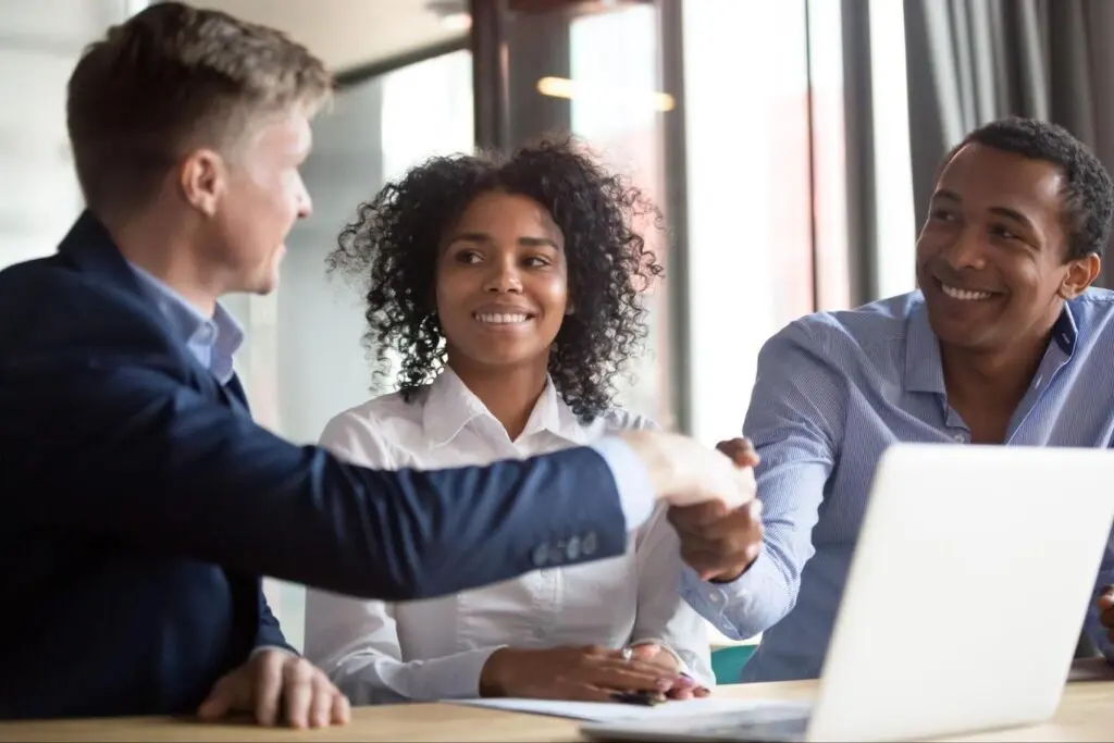 Image of loan officer and two borrowers shaking hands while sitting at a conference table in front of an open laptop.
