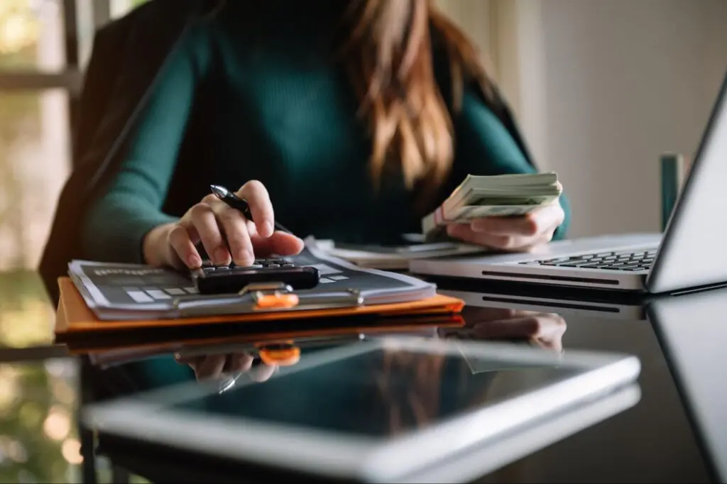 A woman types on a calculator while holding a stack of cash.