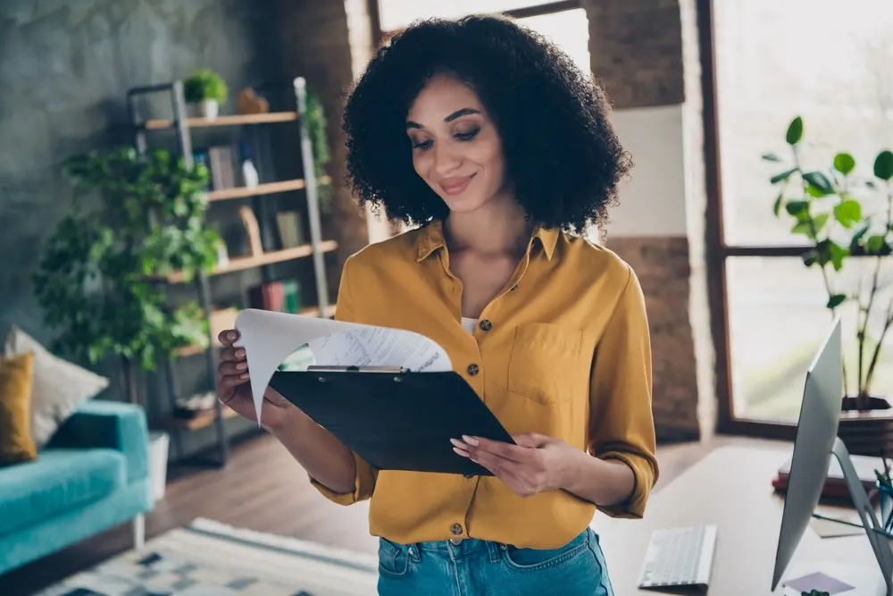 A woman in a yellow button-up and blue jeans stands in her office and looks through documents attached to a clipboard.