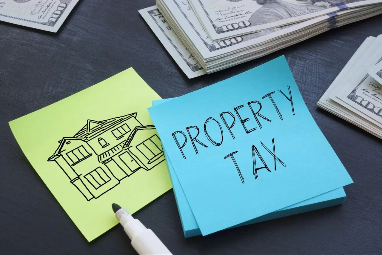 A blue sticky note with “Property Tax” written on it beside a green sticky note with a house drawn on it sitting near stacks of hundred dollar bills.