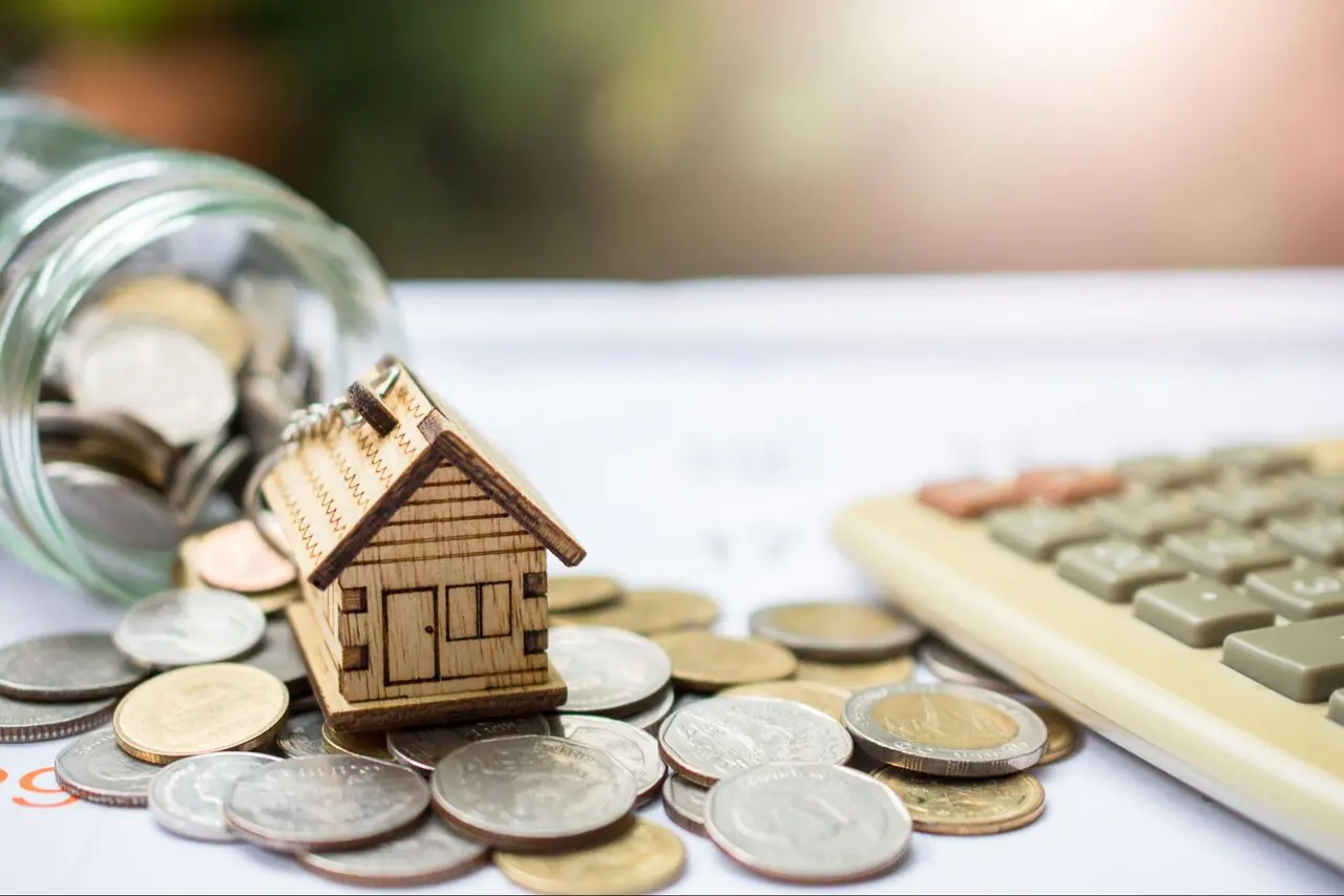 A wooden model home on top of a pile of coins next to a calculator.