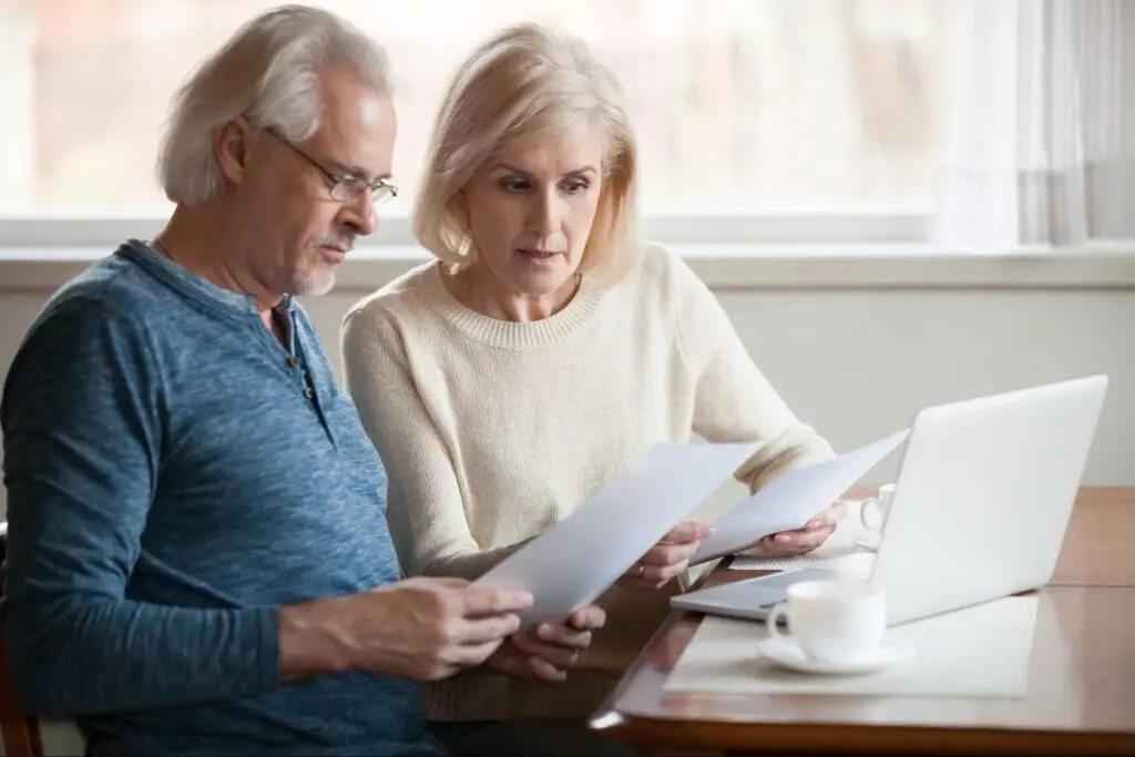 An elderly couple review documents while sitting at a desk with an open laptop in front of them.