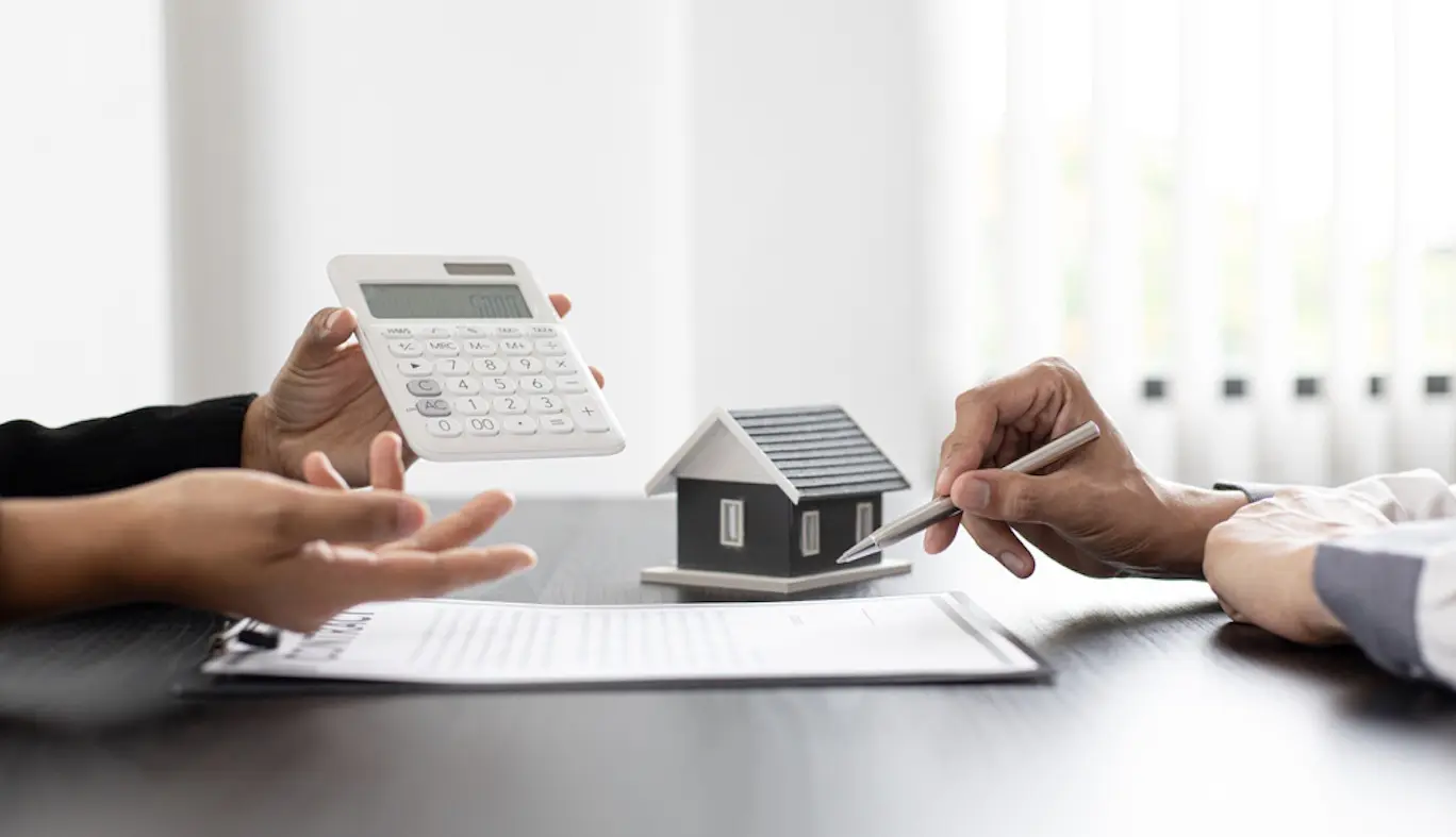 Close up of two people sitting across from each other at a table with one holding a calculator and the other holding a pen that hovers over a document. A small model house is sitting on the table.