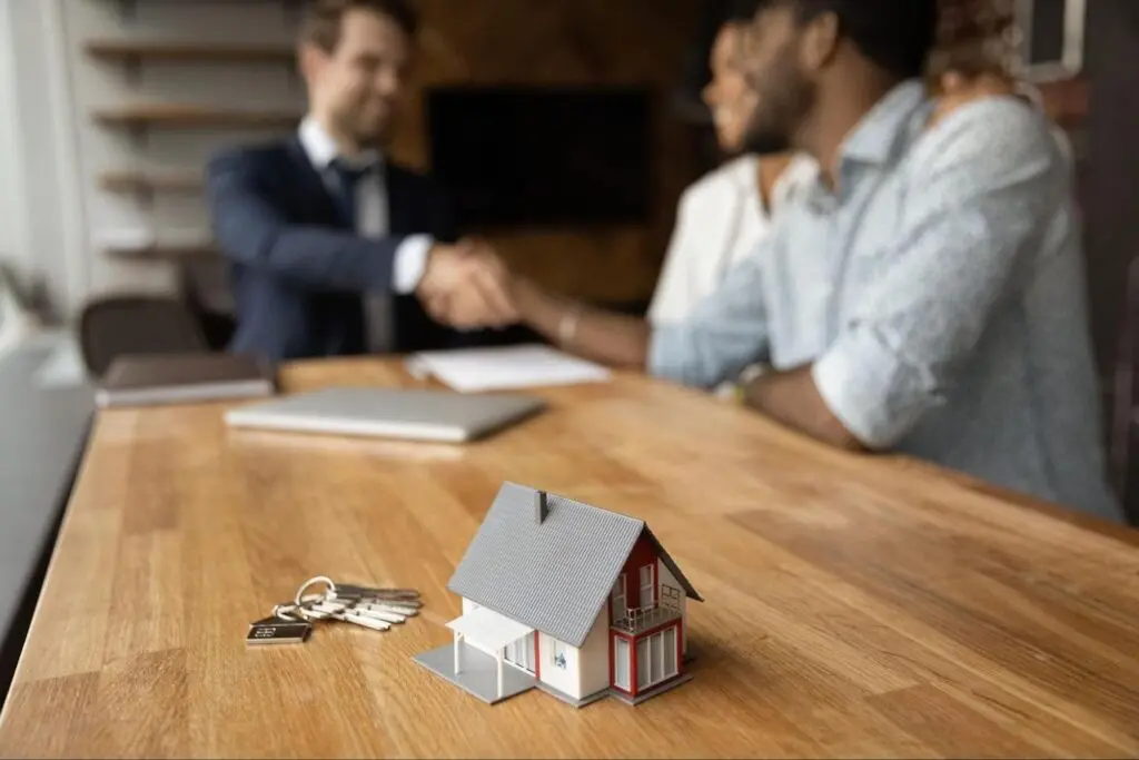 A model home and set of keys sit on a desk in the foreground while in the background a real estate agent wearing a suit shakes hands with a couple.